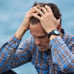 A man in a plaid shirt sits by the water looking distressed, symbolizing stress.